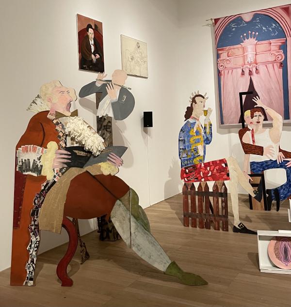 a corner of the gallery shows big cutouts of people in different poses, singing, playing a flute, sipping a cup, and hugging, all painted in sectioned bits of texture and flat colour, alongside some paintings on the wall of a portrait of a man and a big pink curtain under an arch
