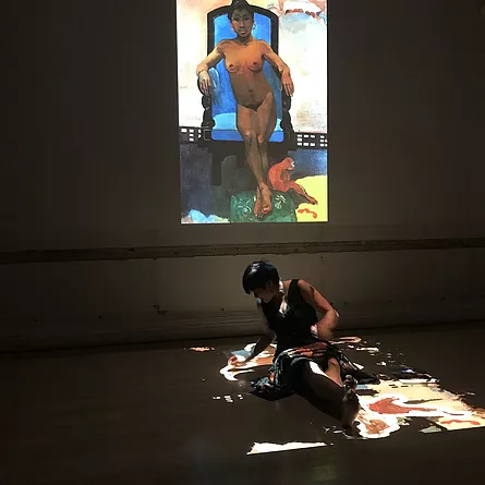 performer is still below the projection, but now there&rsquo;s a painting of a nude brown woman sitting with her arms either side of her on a bright blue chair