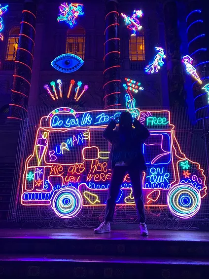 Zarina stands in front of a group of neons, namely a huge neon of an ice cream truck full of colour and shapes and swirls and stars