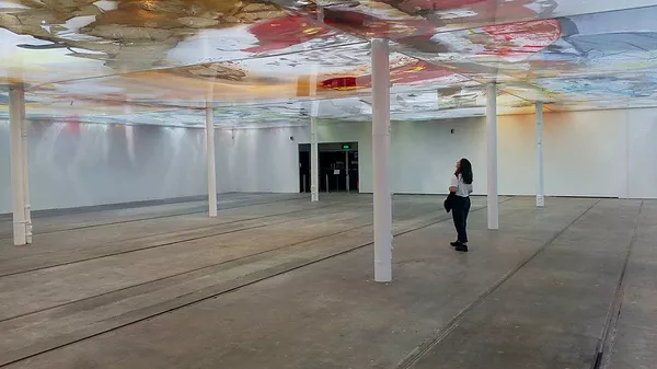 Gabrielle stands in a huge open gallery space broken up with white columns that are holding up big pieces of plastic on top of which the artist has pooled colour and texture in different shapes, so it makes a new colourful ceiling much lower than the gallery’s original one