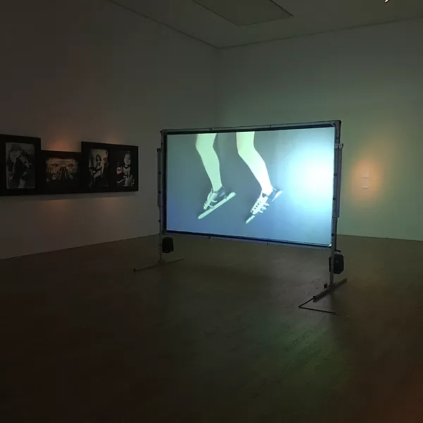 the artists legs hang down from the back of a car, and she is wearing ice skates that scrape against the road as the car moves, and the video is being shown on a projection screen in the middle of a dark gallery space