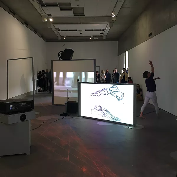 a shot in a big white cube gallery space where a dancer moves between a group fo different projections while a crowd at the back watches