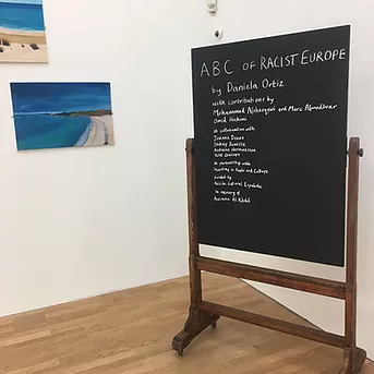 A black board in the corner of the room says the ABC of Racist Europe by Daniela Ortiz