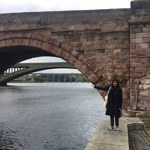 zarina smiles and waves an arm out over the big river she is standing next to, in front of multiple bridges