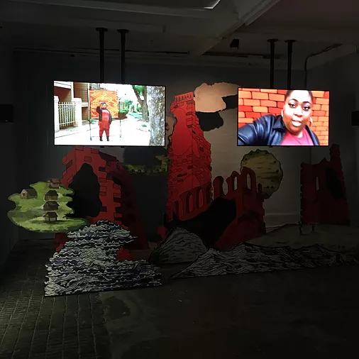 a cardboard cut out set piece of a field with houses, waves, and crumbling towers in bright colours and a cloud, and above, there are two screens on mounts coming down from the ceiling. One shows someone standing in a street, and the other is a black woman holding the camera at a selfie angle