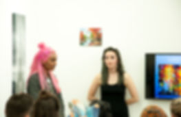 gabrielle and zarina are at the front of the talk, gabrielle has her hands behind her back and zarina is speaking - she also has neon pink hair