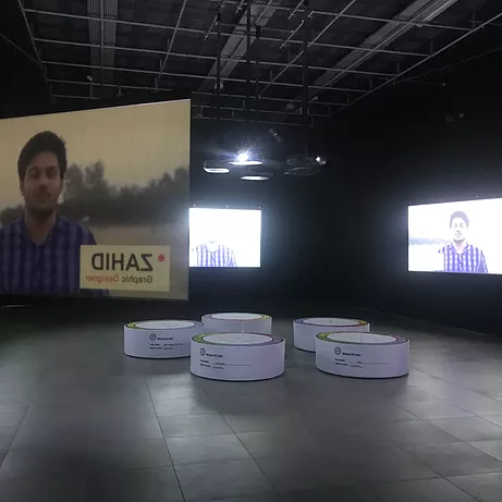 five circular white pie chart sculptures in the centre of a darkened gallery space at at the centre of hanging projections that currently show a man being introduced as Zahid