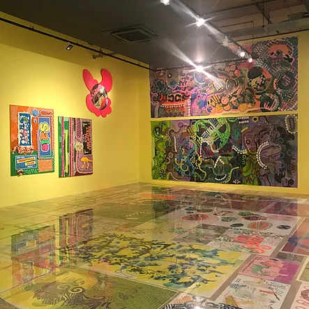 a yellow gallery space has massive abstract pattern colourful paintings on one wall but the same style also continues on the floor as well, except the floor artworks are protected with clear plastic so people can walk over top