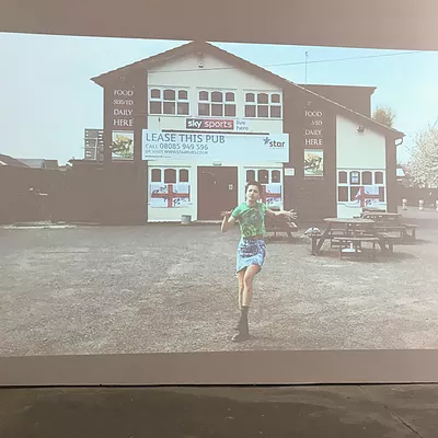 an image of rene matic in a green top and a denim skirt and black boots raising a leg mid dance outside a huge pub with outdoor seating