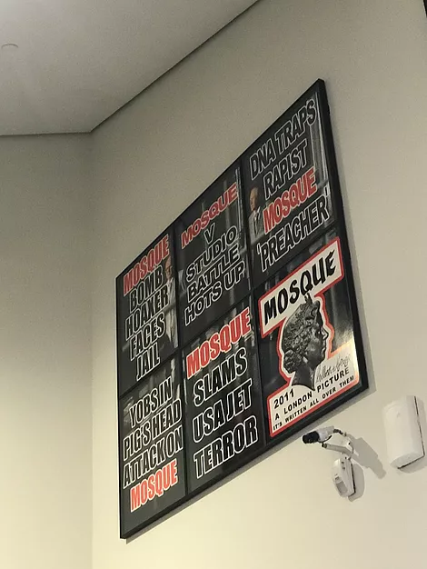 6 framed posters next to one another high up on the wall above a security camera. They are all in a newspaper style, saying things like &lsquo;mosque slams USA jet terror&rsquo; and &lsquo;dna traps rapist mosque preacher&rsquo; and &lsquo;mosque versus studio battle hots up&rsquo;