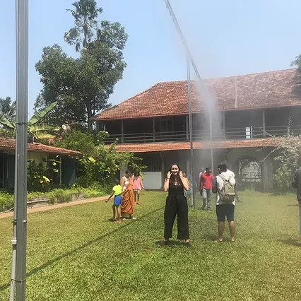 Gabrielle stands in and amongst other people on a field, and everyone is standing under a table metal frame as it shoots out water vapour into the sun