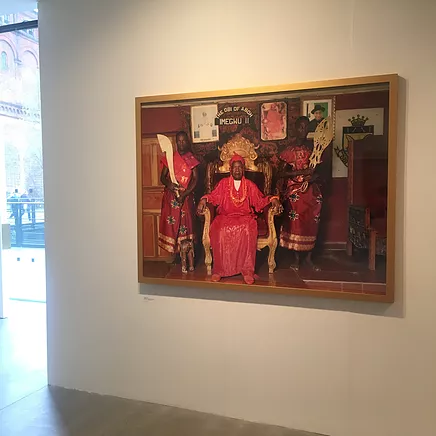 three Black people stand together with the person in the centre on a gold chair like a thrown, and everyone is wearing red and gold