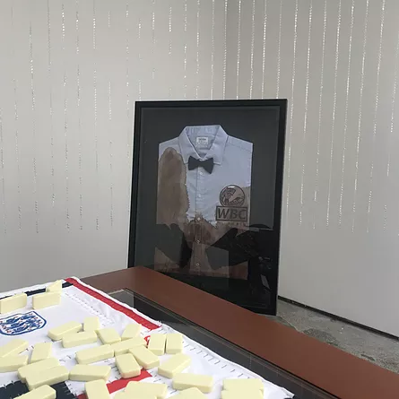 in the corner of the room, a framed white shirt with a WBC logo and a bowtie is stained with something dark brown that could be dirt or blood, and there&rsquo;s england&rsquo;s teams logo with domino pieces upside down and over top