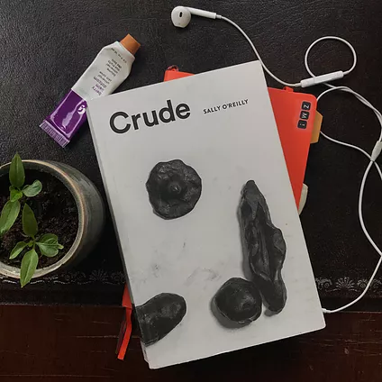 the crude book is on a flatlay, on top of an orange notebook, some headphones, next to a glossier lip balm, and a tiny plant. the book cover itself has some black burnt out sculptures that look like fruit or veg, or more suggestive, like the open pea pod could be labia instead