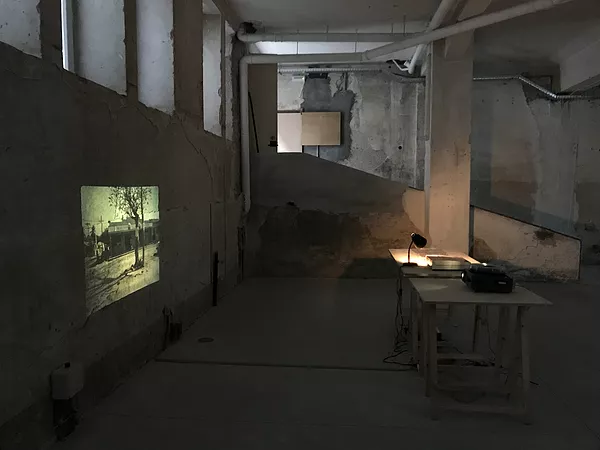 a rough cement underground gallery space has a slide carousel on a desk casting out an old image of a tree in front of a building