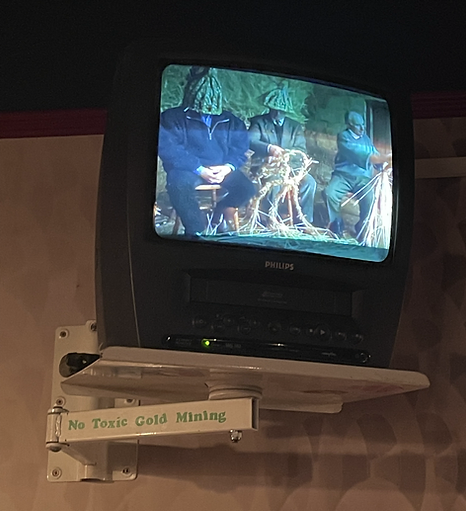 A CRT screen shows three people sat down making something, and two of them have their heads completely covered by green woolly hats. There is a decal below the tv on the tv stands that says no toxic gold mining