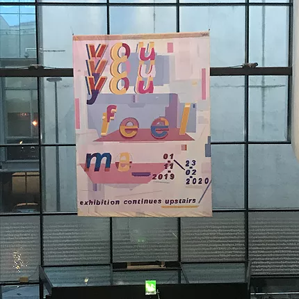 an exhibition poster hanging in the lobby, says you feel me with a pastel colourful abstract graphic