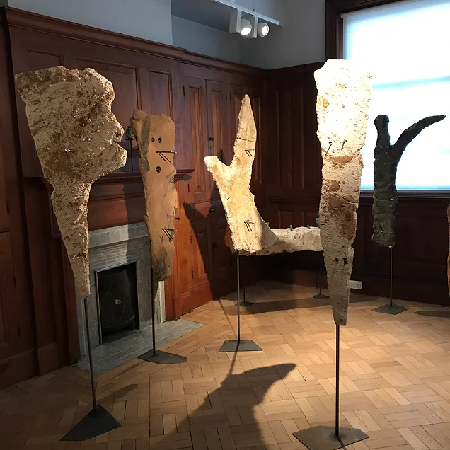 large sculptures on metal stands are arranged around a wood panelled room. they sort of look like ripped pieces of cardboard but huge, the height of people, an some have cut out shapes like arms or metal and holes where faces might be, but totally abstract