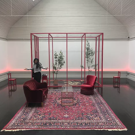 the gallery space is furnished like a little showroom with a big carpet, couch, armchair, and then a tall red framed cage with birds inside