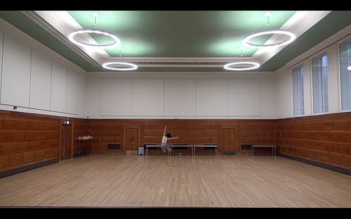 a wide screen shot of a big hall with wooden floors and big circular lights on the ceiling while Rene is at the back of the space jumping in the air with arms up and out