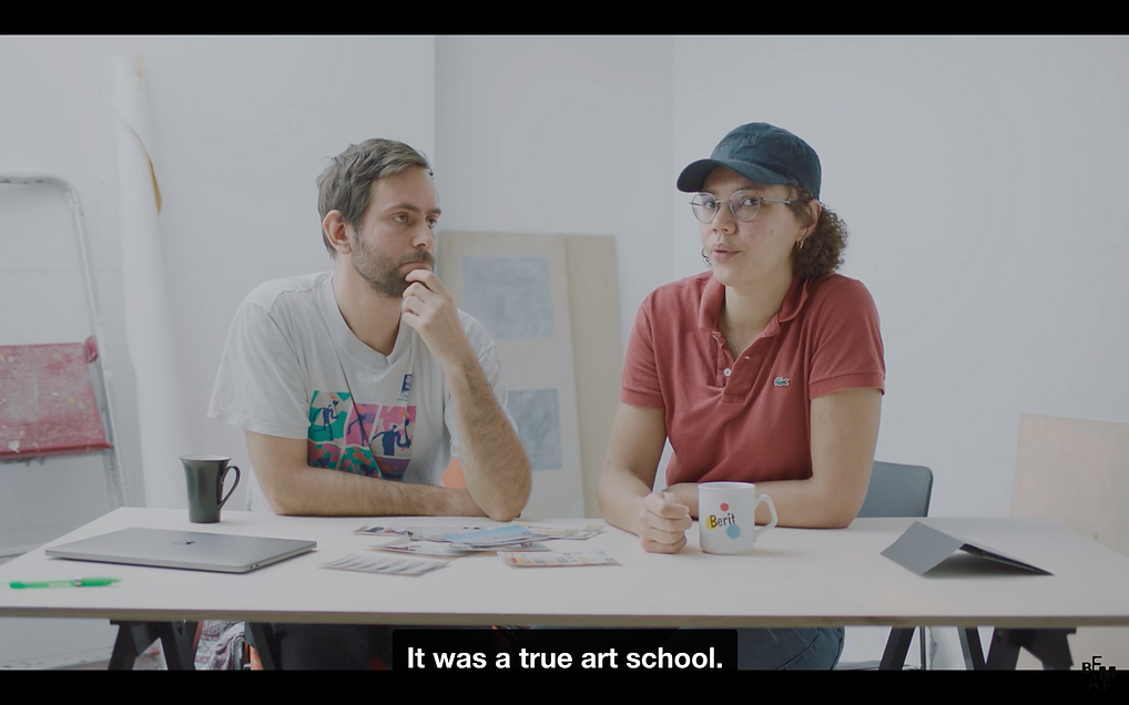 two white peple sit at a desk in a studio with laptops, mugs, and the caption says it was a true art school