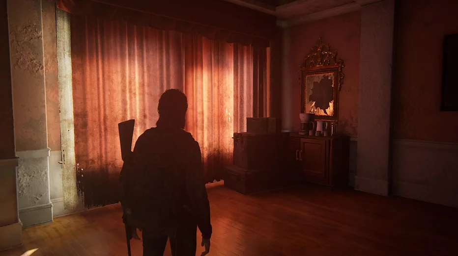 Ellie stands in a room with a rifle on her back. The red curtains are closed and the room is glowing softly with light. We can see boxes and a broken ornate mirror but apart from that it is empty