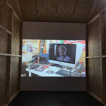 a projection in a tiny chipboard theatre shows a macbook on a table with an old woman on the screen in a bonnet