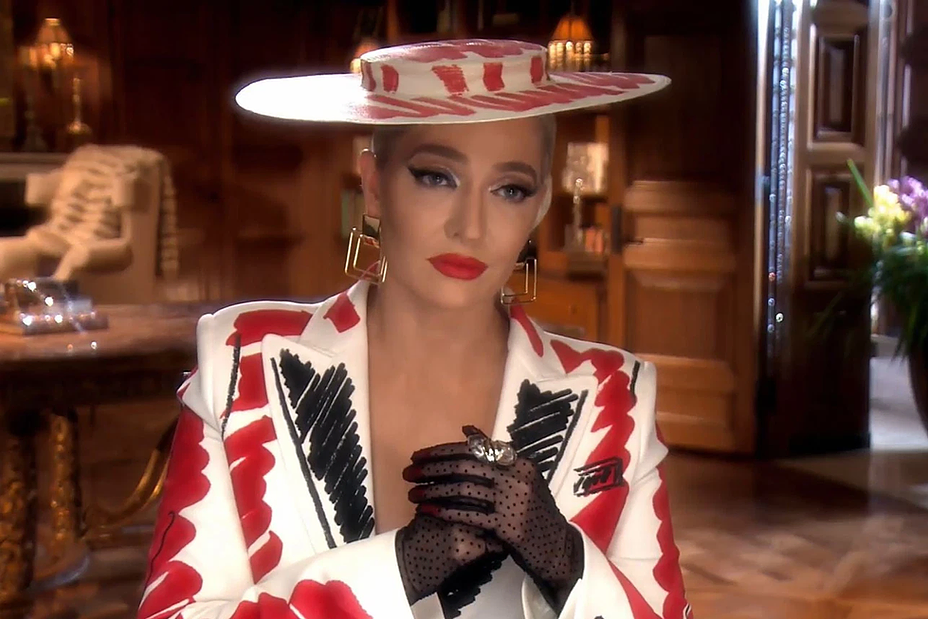 Erika Jayne wears an absolutely insane suit jacket and thin hat, all white, except there is a black and red print in top that looks like someone has scribbled the colour and shape into the outfit